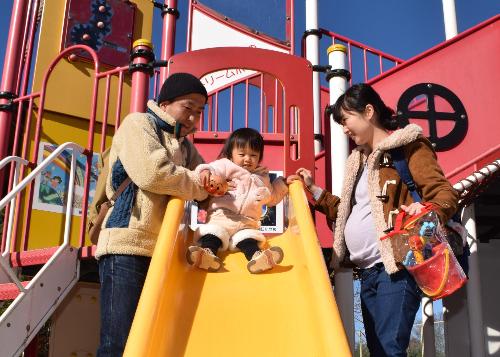 桶川市内の公園の遊具で遊ぶ親子の画像。母親が見守るなか、父親が子どもを支えながら滑り台を滑らそうとしている。