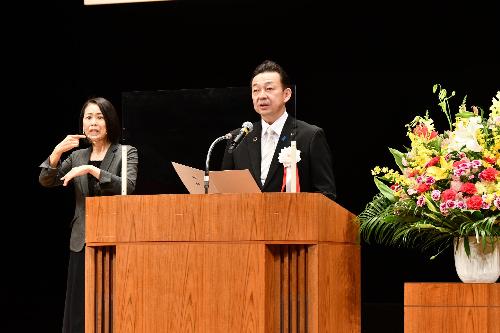 桶川市市制施行50周年記念式典で式辞を述べる小野桶川市長画像。市長の左となりに手話通訳者が並ぶ。