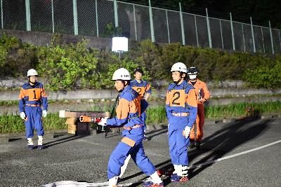 埼玉県操法大会に出場するため訓練を重ねる桶川市消防団の様子の画像。消防職員から操法の所作について指導を受ける4人の団員が写っている。中央の団員は消防ホースを構え、消火の所作を見せている。