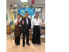 左から、クイーンズビラ女性職員、桶川市長、神頭施設長の3人が桶川栄養まつりの横断幕の前でピースをしている写真