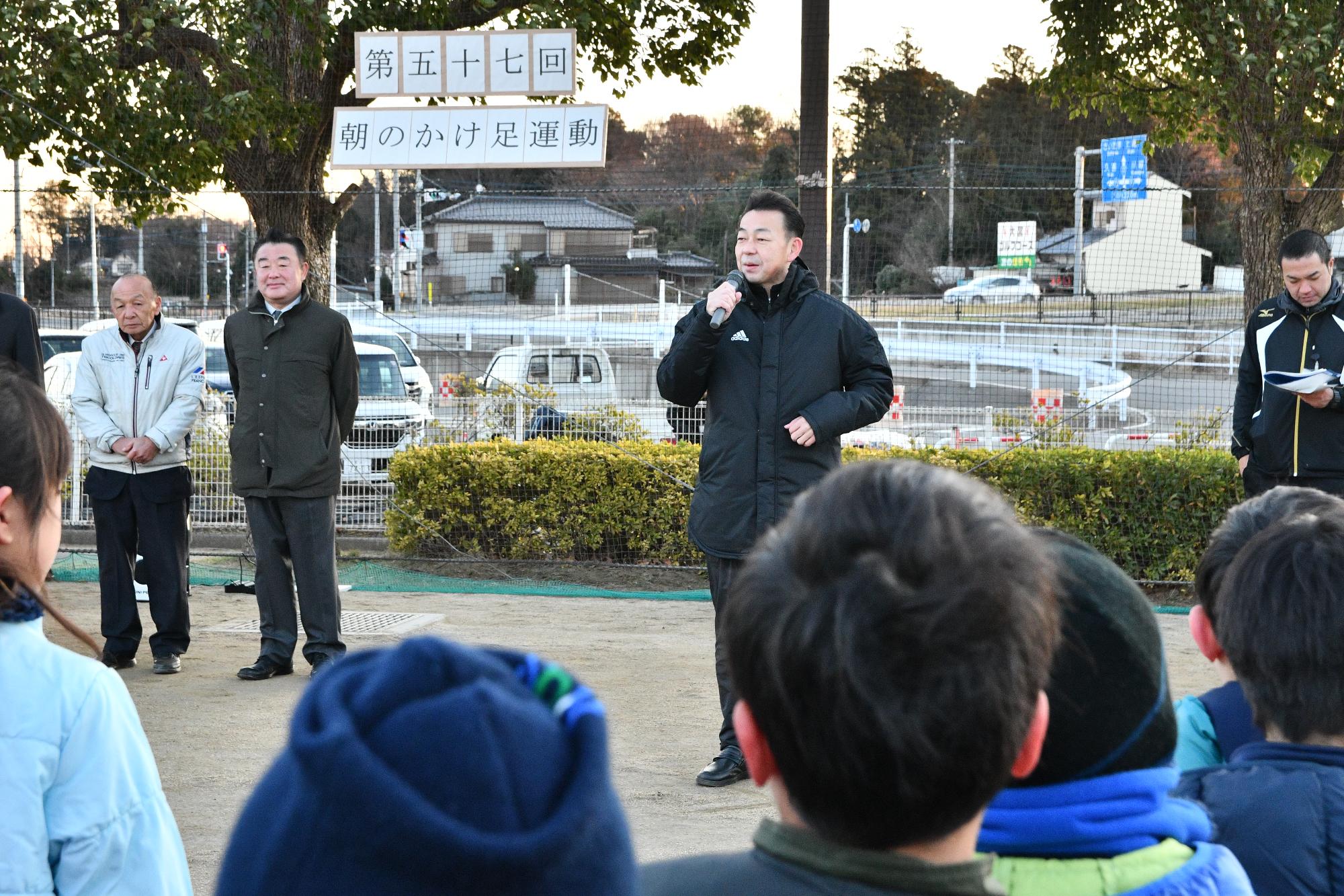 朝のかけあし運動の城山公園にて、大勢の子どもたちの前で市長が挨拶をしている